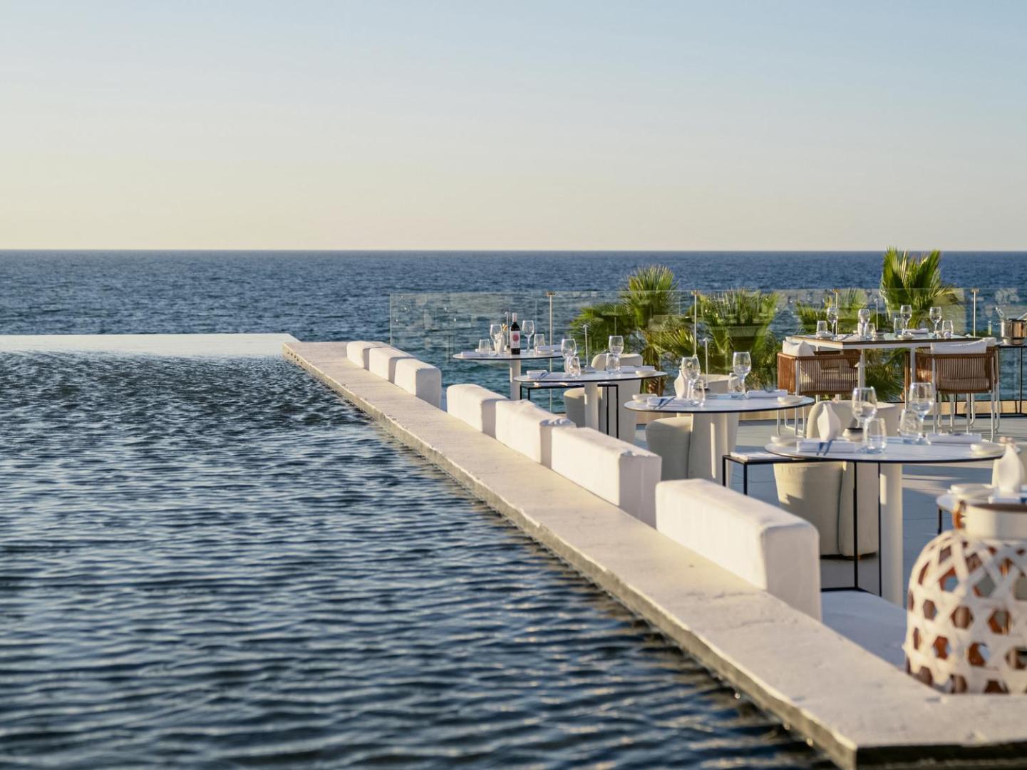 Grecotel Lux Me White Palace restorāna terase pie bezgalības baseina ar galdiņiem un skatu uz jūru — eleganta vide vakariņām