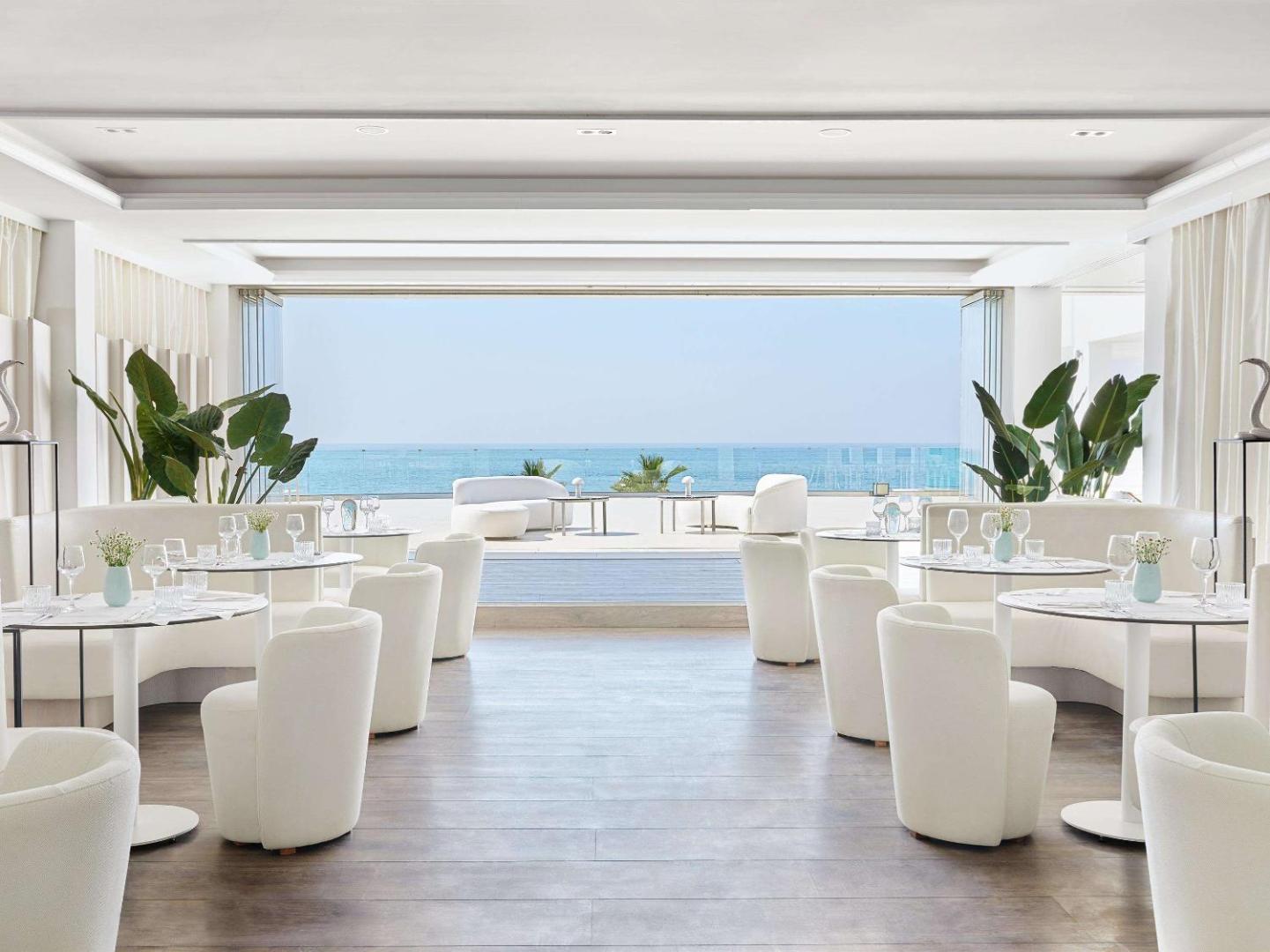 Grecotel Lux Me White Palace gaišais un modernais restorāns ar panorāmas logiem uz jūru — balts interjers un eleganti mēbelēta zāle
