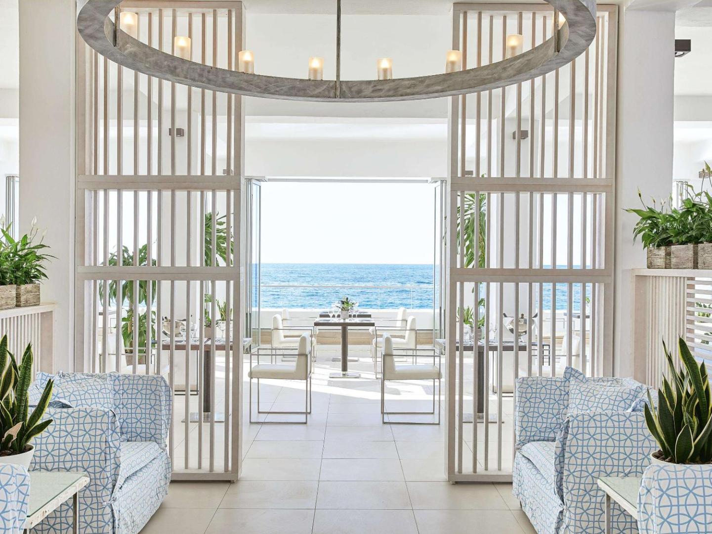 Grecotel Lux Me White Palace restorāna interjers ar gaišām mēbelēm un panorāmas skatu uz Vidusjūru caur atvērtām durvīm