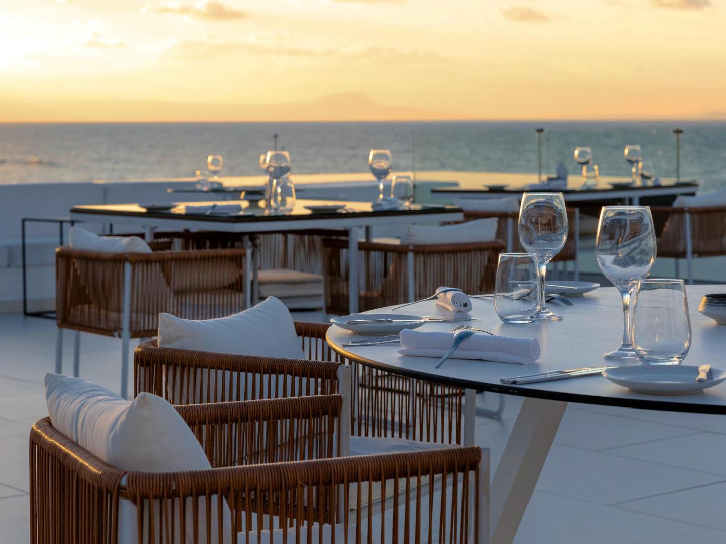 Grecotel Lux Me White Palace restorāna galdiņu klājums saulrieta gaismā uz terases — skats uz jūru un kalniem tālumā