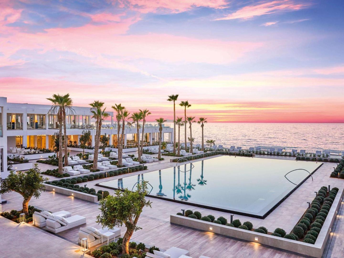 Grecotel Lux Me White Palace viesnīcas panorāma saulrietā — izgaismots baseins, palmas un rozā debesis virs jūras Krētā