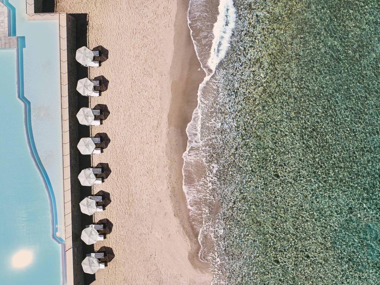 Aerofoto skats uz Grecotel Lux Me White Palace pludmali ar saulessargiem, smilšu joslu un dzidru, tirkīzzilu jūras ūdeni