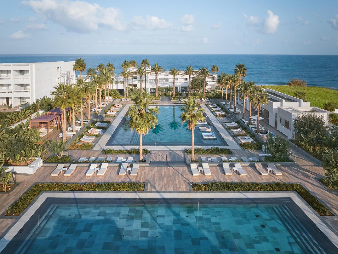 Grecotel Lux Me White Palace plašā baseinu zona ar palmām un skatu uz jūru Krētā — sauļošanās krēsli un moderna arhitektūra