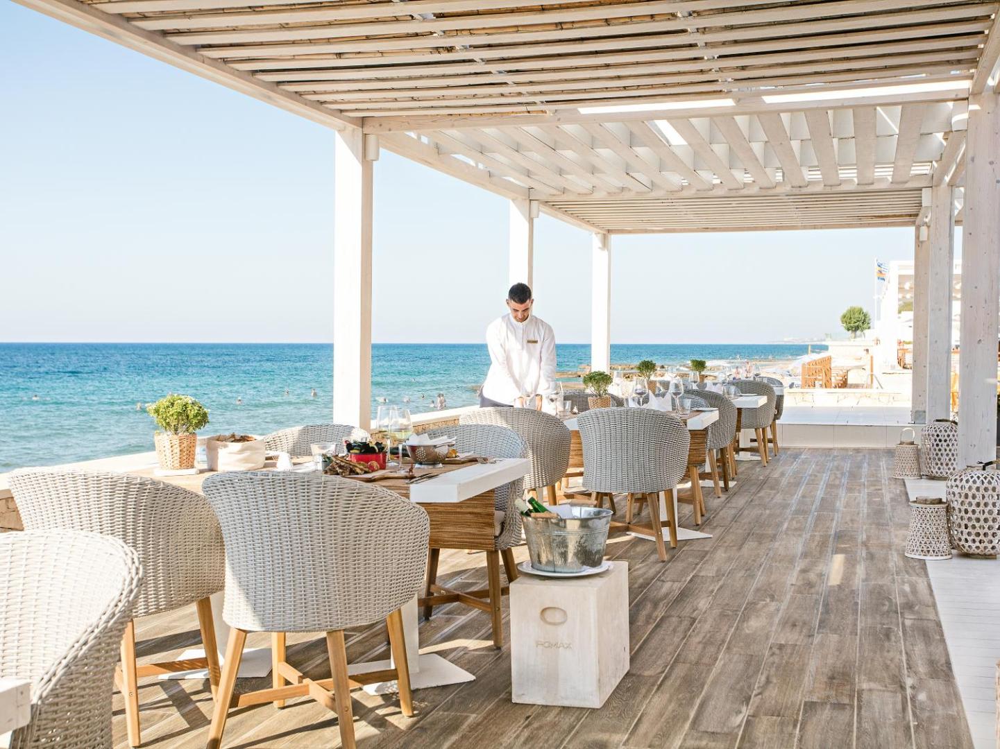 Grecotel Lux Me White Palace restorāna terase ar skatu uz jūru, baltām mēbelēm un eleganti klātiem galdiem Krētas piekrastē