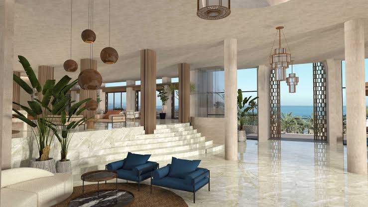 Nana Royal Premium Resort lobby atpūtas zona ar zilām mēbelēm, marmora kāpnēm un skatu uz jūru caur lieliem logiem.