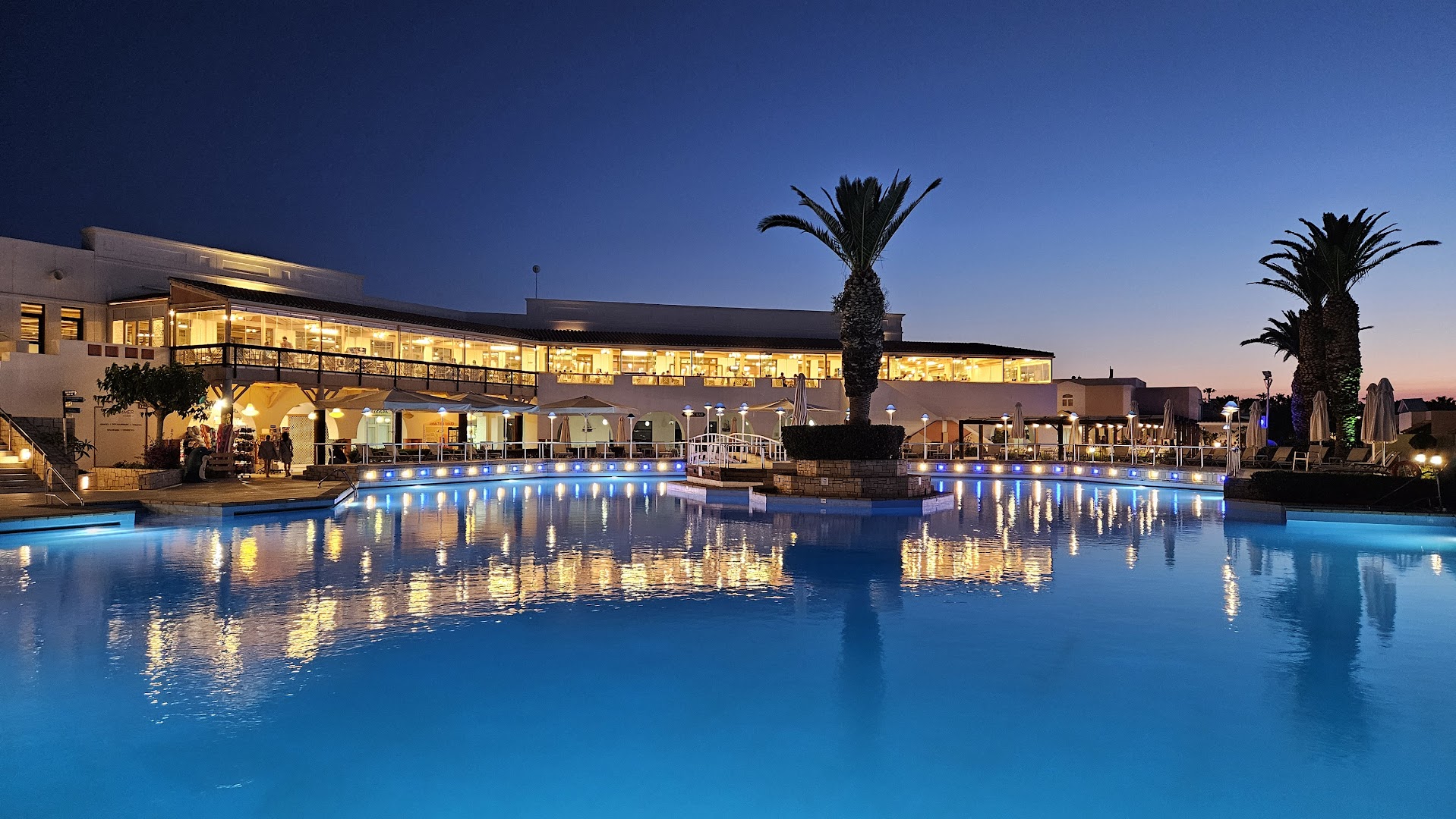 Nana Royal Premium Resort galvenā ēka un izgaismots baseins vakarā — nakts apgaismojums atspīd ūdenī pie restorāna terases