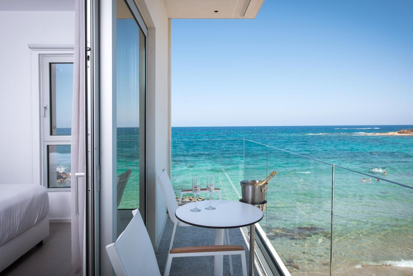 Enorme Ammos Beach Resort balkons ar stikla margām, skatu uz tirkīzzilo jūru un dzirkstošo vīnu Krētā