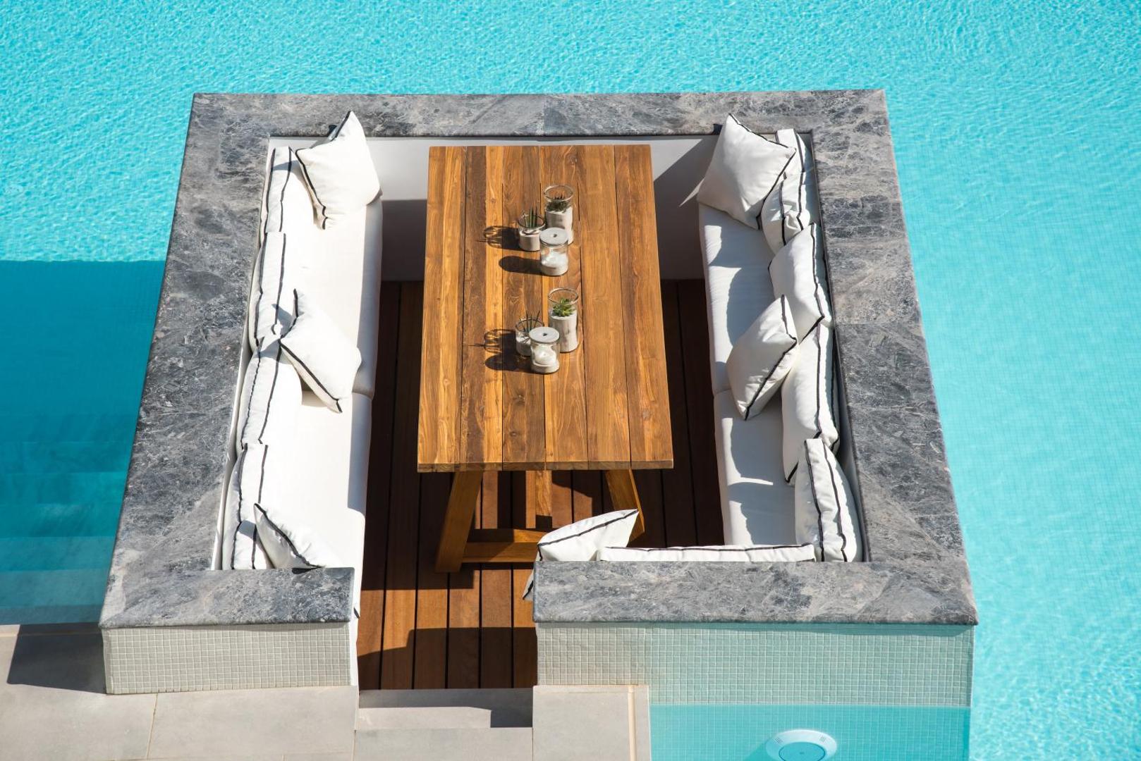 Tuvplāns Enorme Ammos Beach Resort baseina atpūtas zonai ar koka galdu, baltiem spilveniem un pelēku akmens apdari