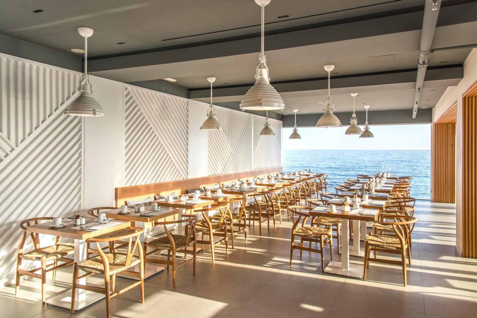 Enorme Ammos Beach Resort modernais restorāns ar gaišu interjeru, koka mēbelēm un panorāmas skatu uz Vidusjūru