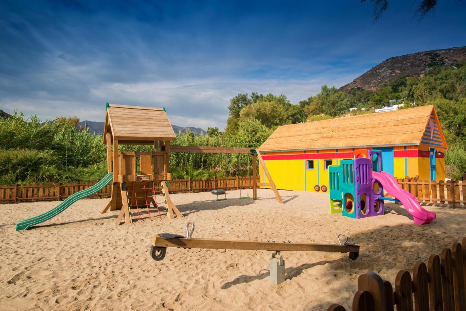 Fodele Beach & Water Park Resort bērnu rotaļu laukums ar smiltīm, slidkalniņiem un šūpolēm Krētā uz kalnu fona