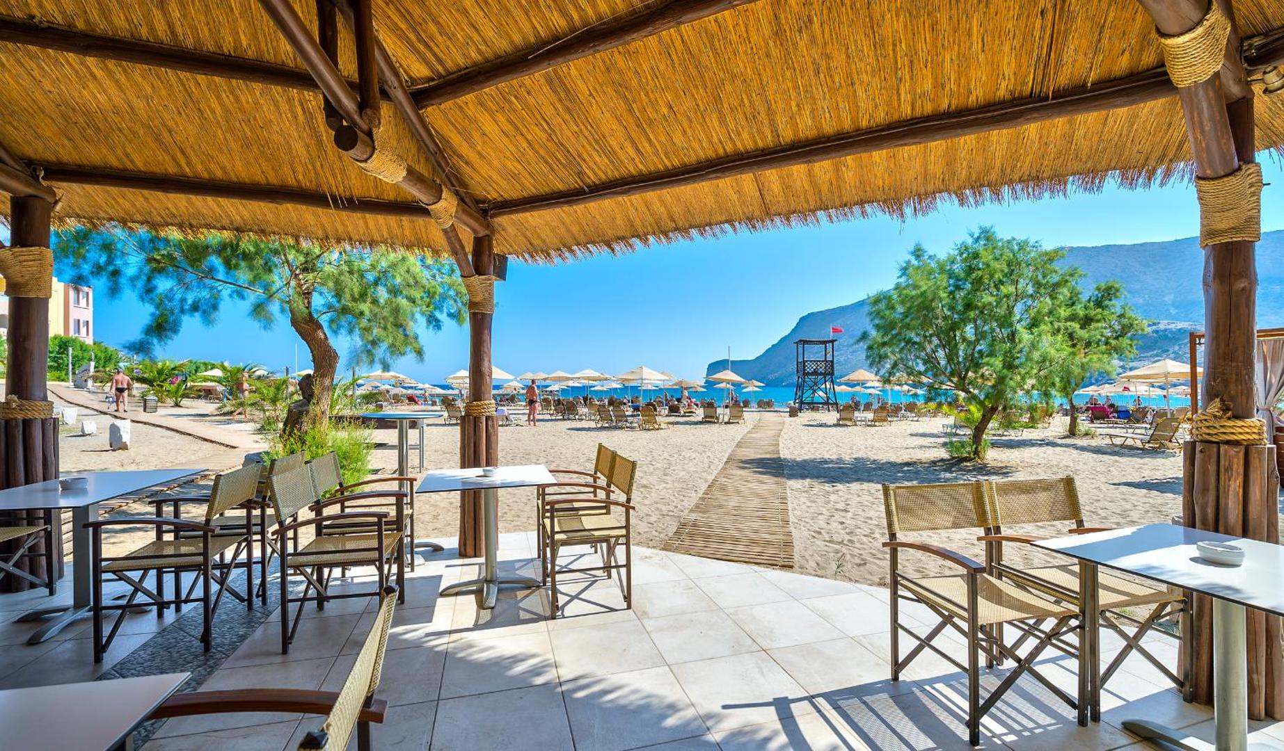 Fodele Beach & Water Park Resort pludmales bārs ar koka nojumi un skatu uz smilšu pludmali, saulessargiem un kalniem tālumā