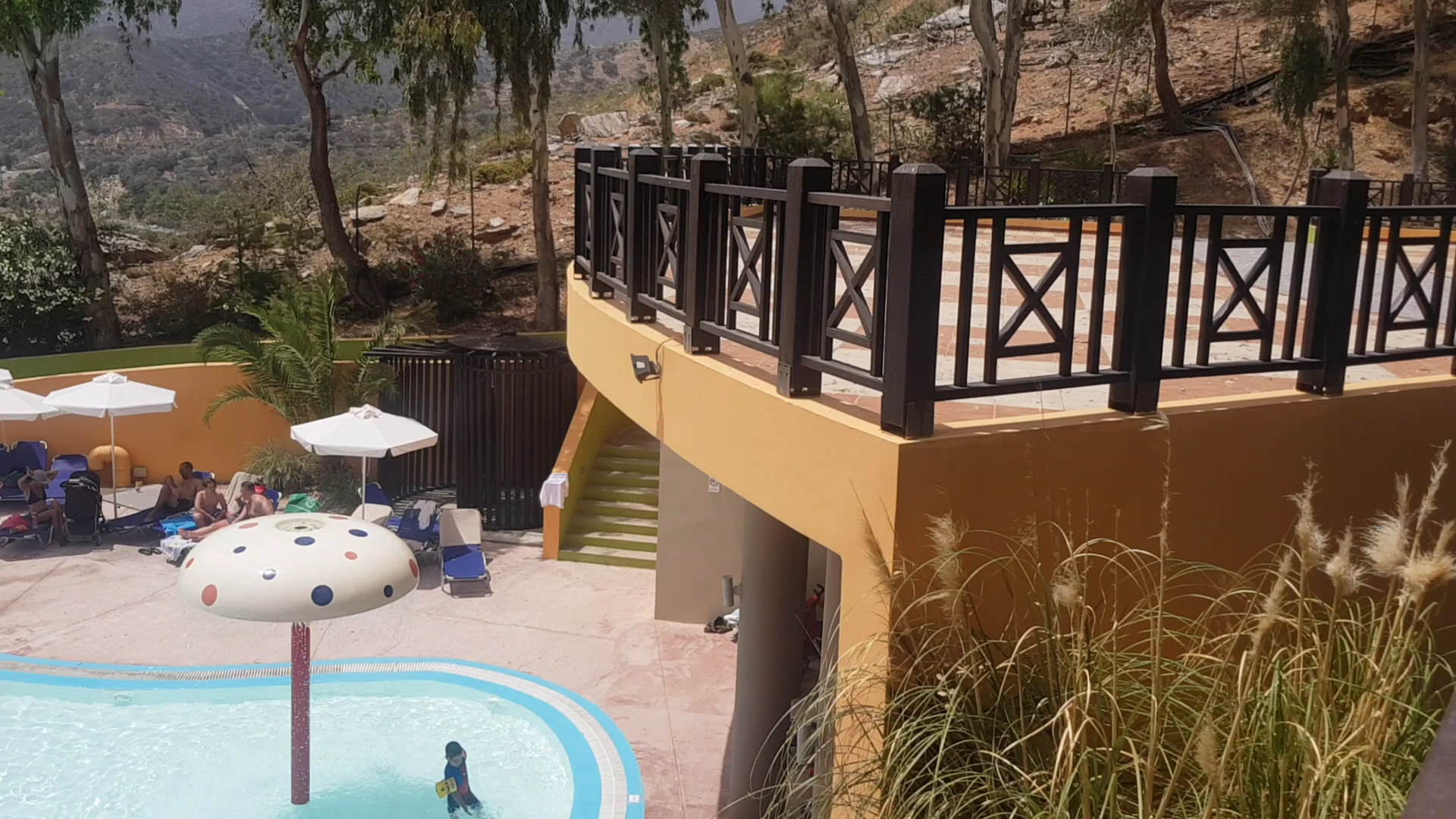 Fodele Beach & Water Park Resort bērnu baseins ar sēnes formas strūklaku, saulessargiem un atpūtas krēsliem pie kalna nogāzes