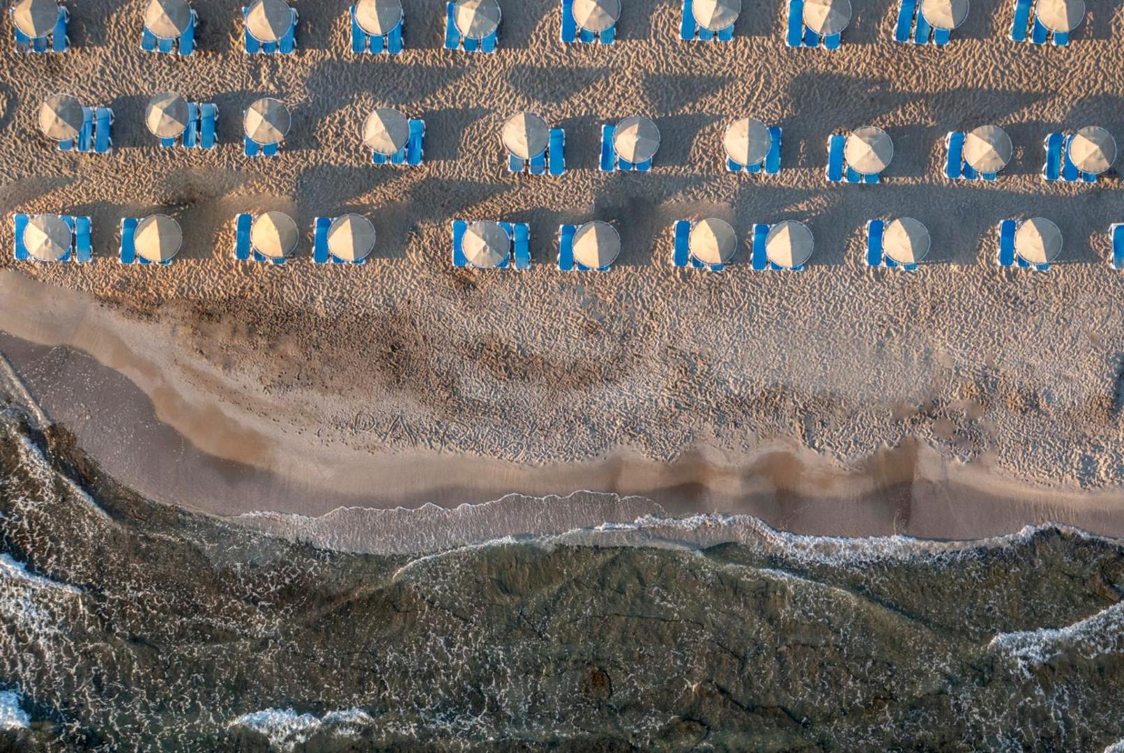 Lyttos Beach privātā pludmale no putna lidojuma — sakoptas saulessargu rindas un zili sauļošanās krēsli pie dzidrā jūras krasta.