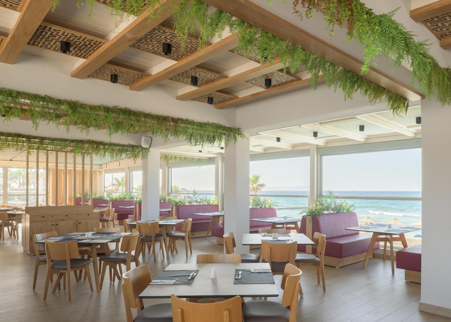 Gaišs Lyttos Beach restorāns ar modernām mēbelēm, zaļumiem pie griestiem un lieliem logiem uz Vidusjūru