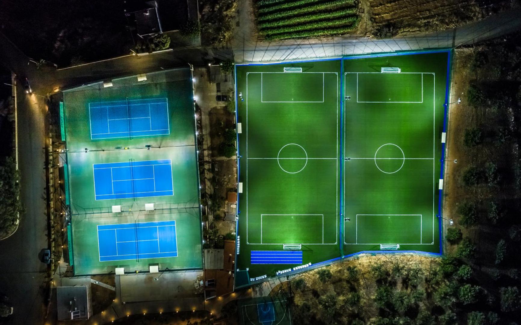 Lyttos Beach sporta kompleksa skats no augšas naktī — apgaismoti tenisa korti un futbola laukumi Hersonisā, Krētā