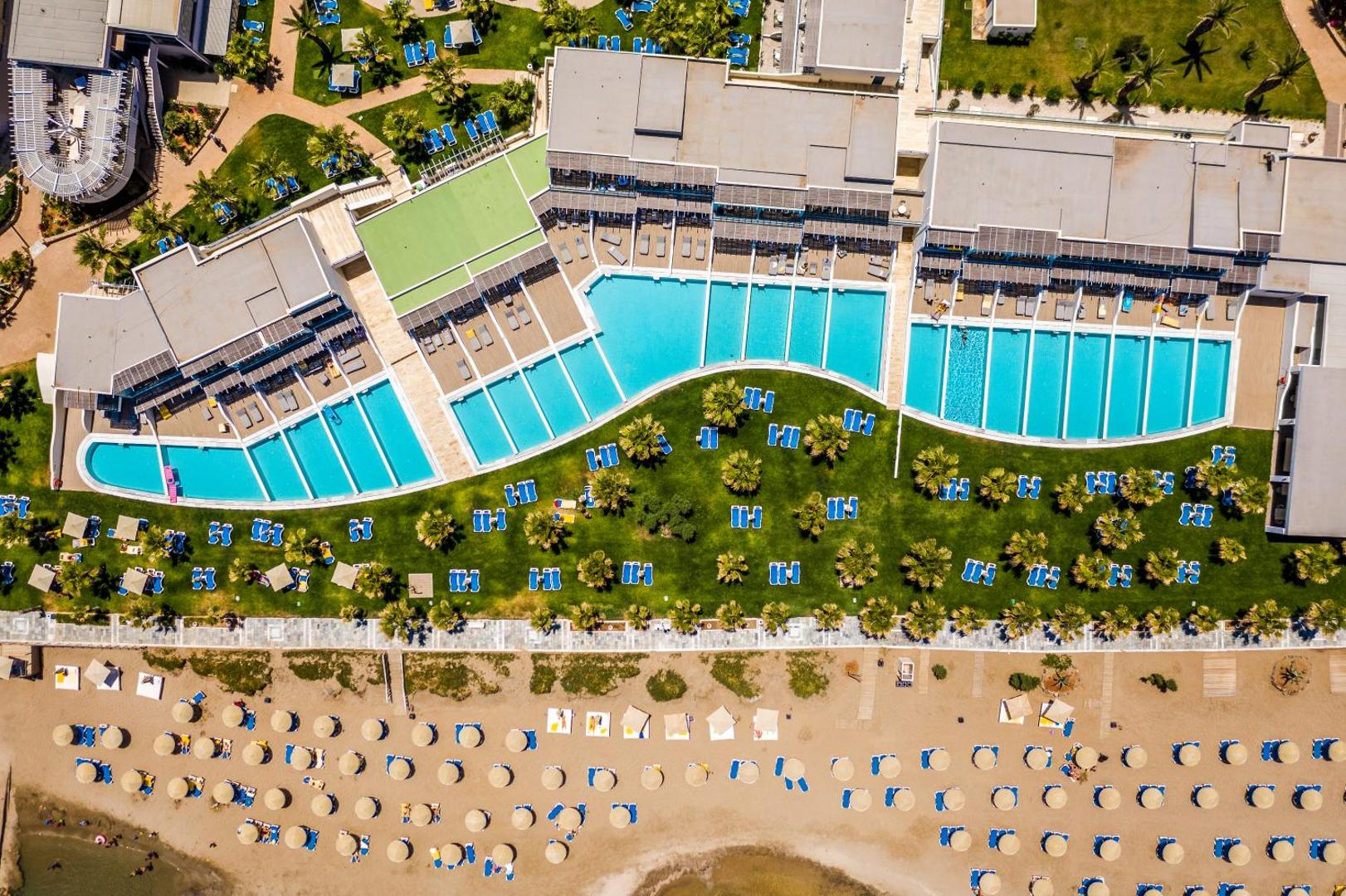 Lyttos Beach viesnīcas skats no augšas uz baseiniem, zaļo zonu un privāto pludmali ar saulessargiem Krētā