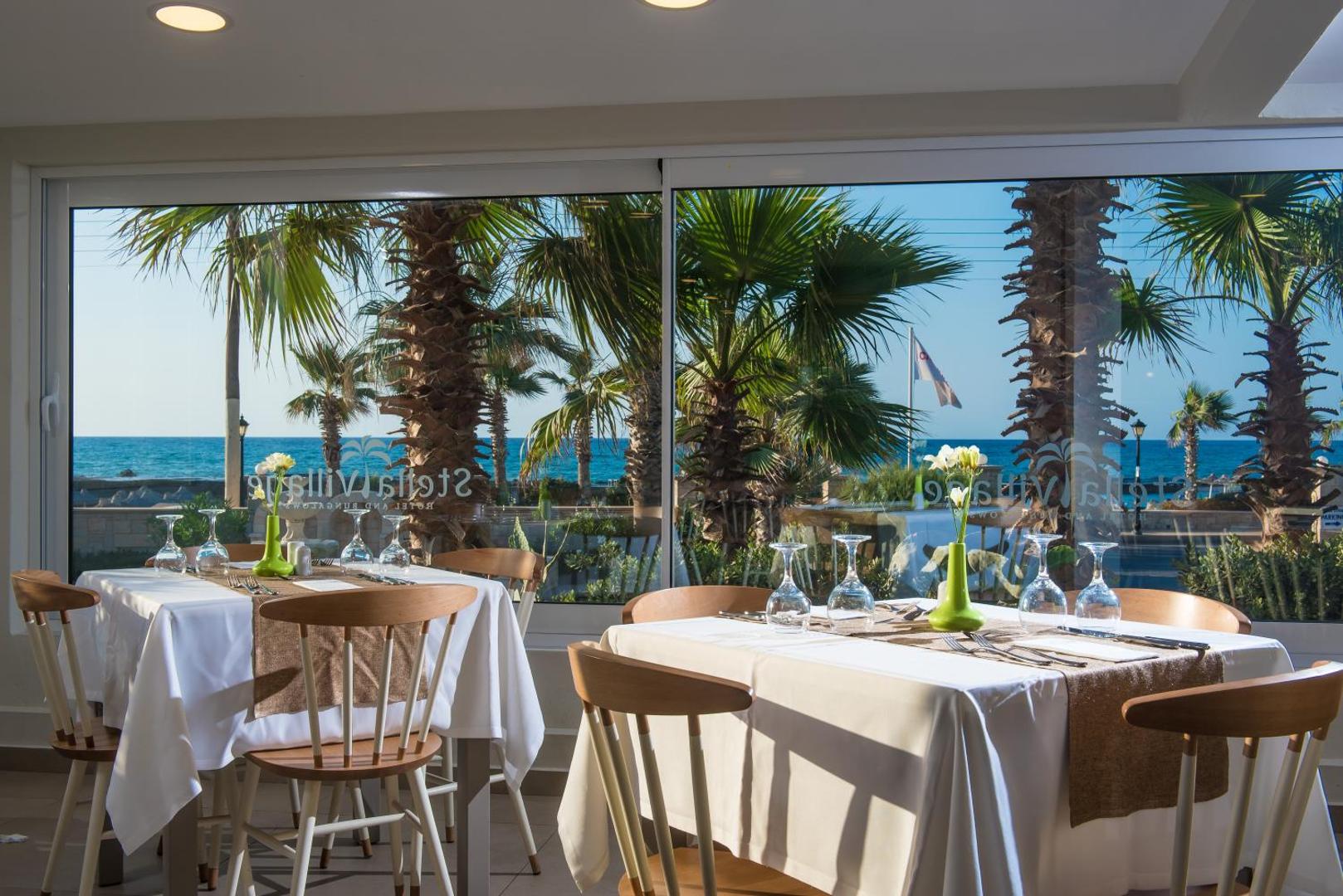 Stella Village Seaside Hotel restorāna galdi ar baltiem galdautiem pie liela loga ar skatu uz palmām un zilo jūru
