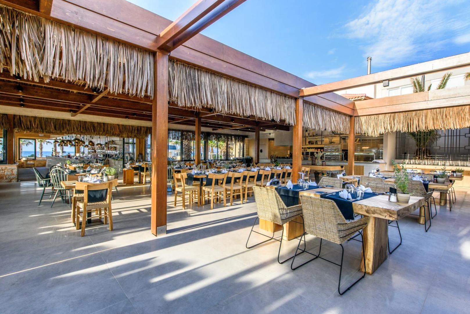 Stella Village Seaside Hotel modernais restorāns ar koka konstrukcijām un salmu dekorācijām, gaišām mēbelēm un plašu āra terasi