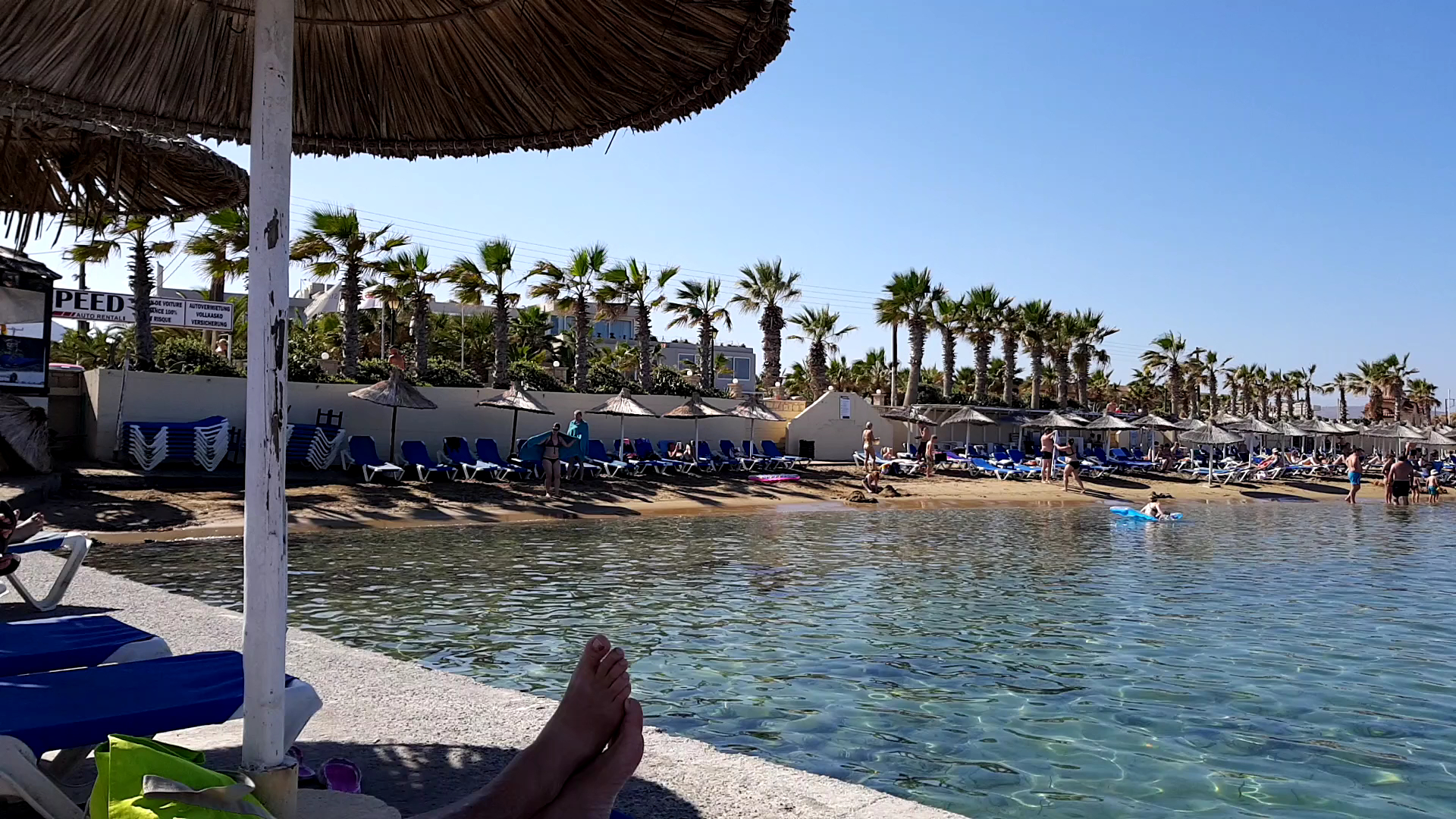 Skats no Stella Village Seaside Hotel pludmales uz dzidro jūras ūdeni, palmām un sauļošanās zonu saulainā dienā Krētā.