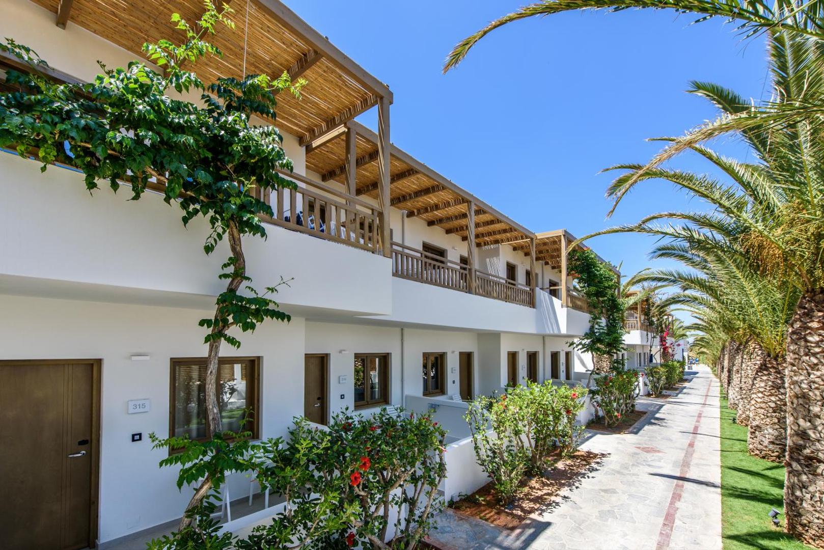 Stella Village Seaside Hotel divstāvu korpusi ar balkoniem un terasēm Hersonisā, Krētā — labiekārtoti gājēju celiņi, palmas un ziedoši krūmi.