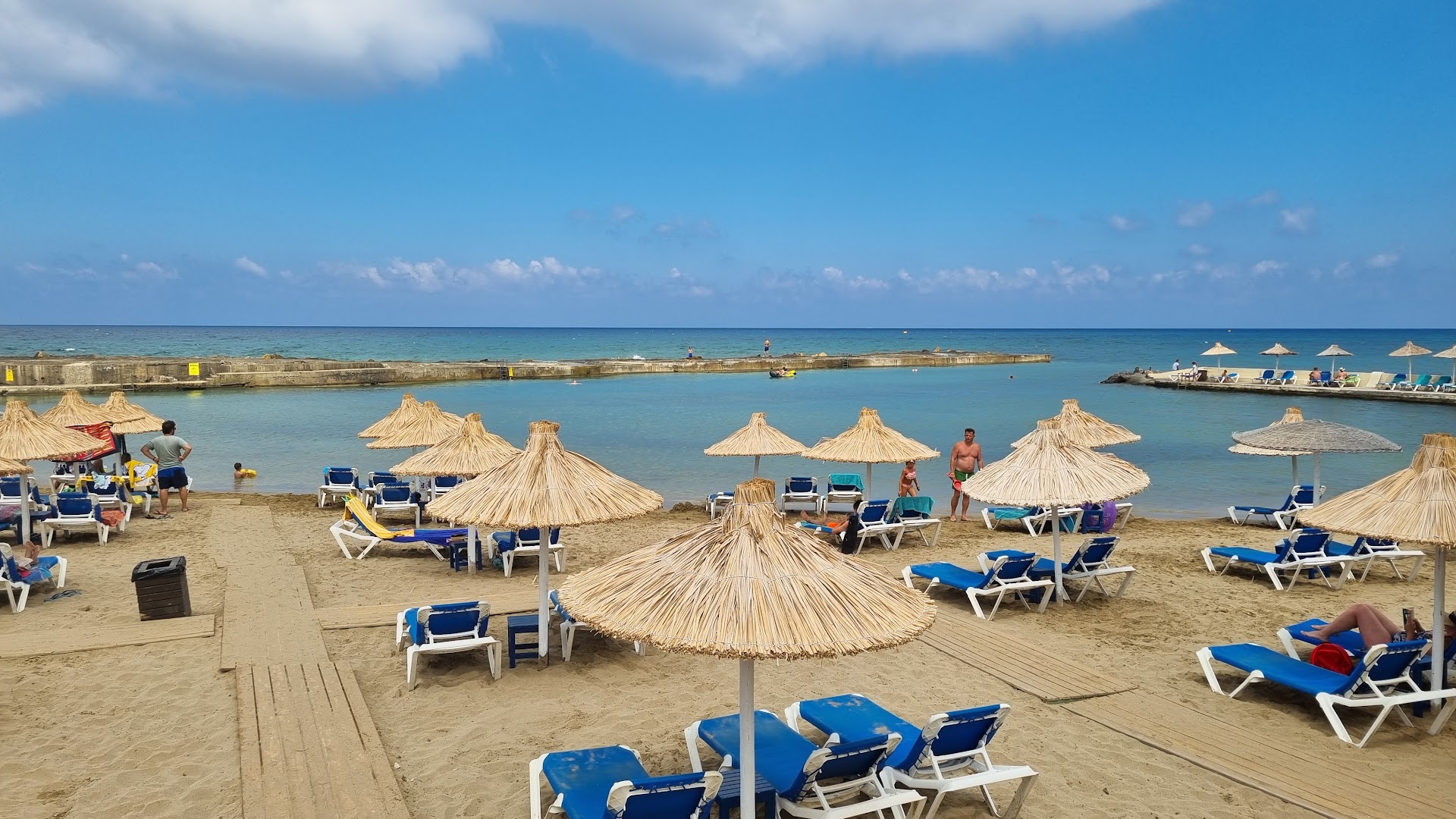 Stella Village Seaside Hotel pludmales zona ar salmu saulessargiem, sauļošanās krēsliem un skatu uz aizsargātu līci un molu Hersonisā.