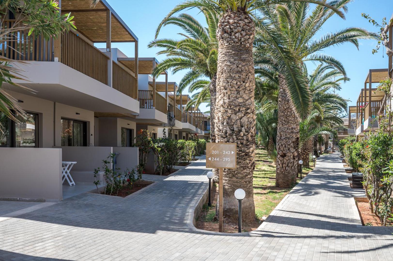 Stella Village Seaside Hotel teritorijas skats ar norāžu zīmi, bruģētiem celiņiem un augstām palmām starp viesnīcas dzīvojamajiem korpusiem.