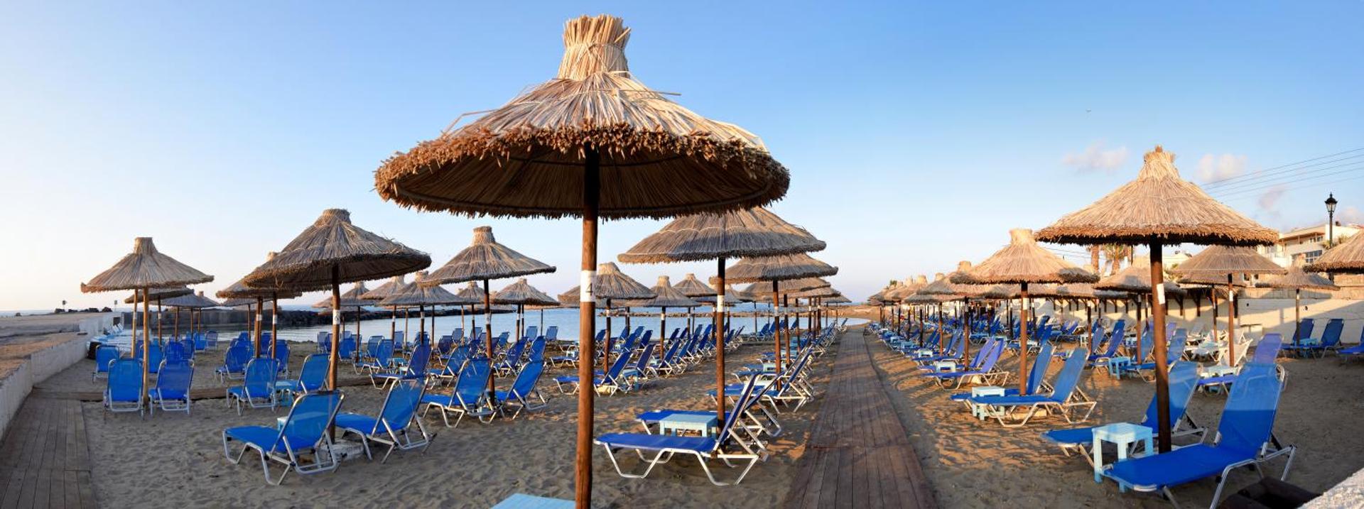 Stella Village Seaside Hotel privātā smilšu pludmale ar daudziem saulessargiem un ziliem sauļošanās krēsliem pie mierīgas jūras Krētas piekrastē.