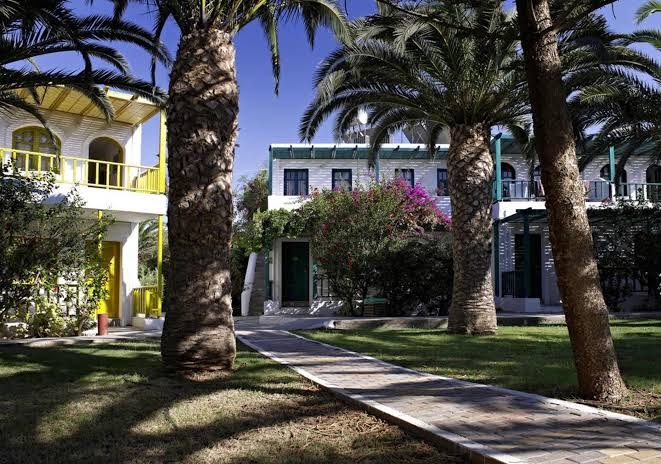 Stella Village Seaside Hotel dārza zona ar zālienu, bruģētu taciņu un palmu ēnām, fonā redzami viesnīcas korpusi ar dzelteniem akcentiem.