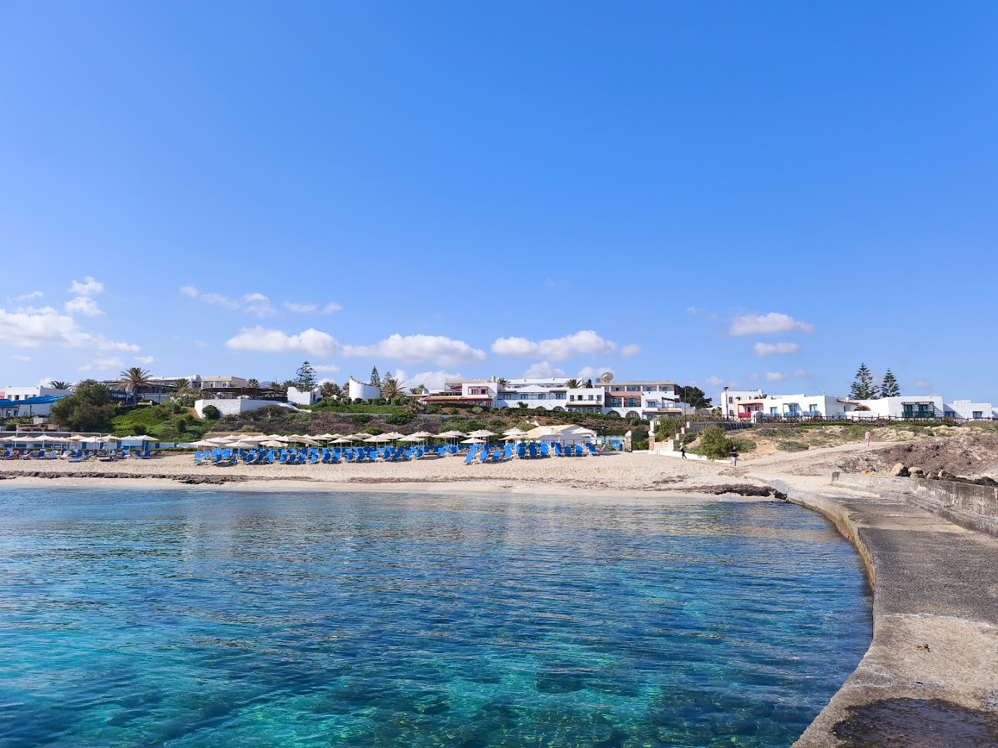 Skats no jūras uz Canvas by Mitsis Cretan Village privāto pludmali ar ziliem saulessargiem un baltām viesnīcas ēkām piekrastē