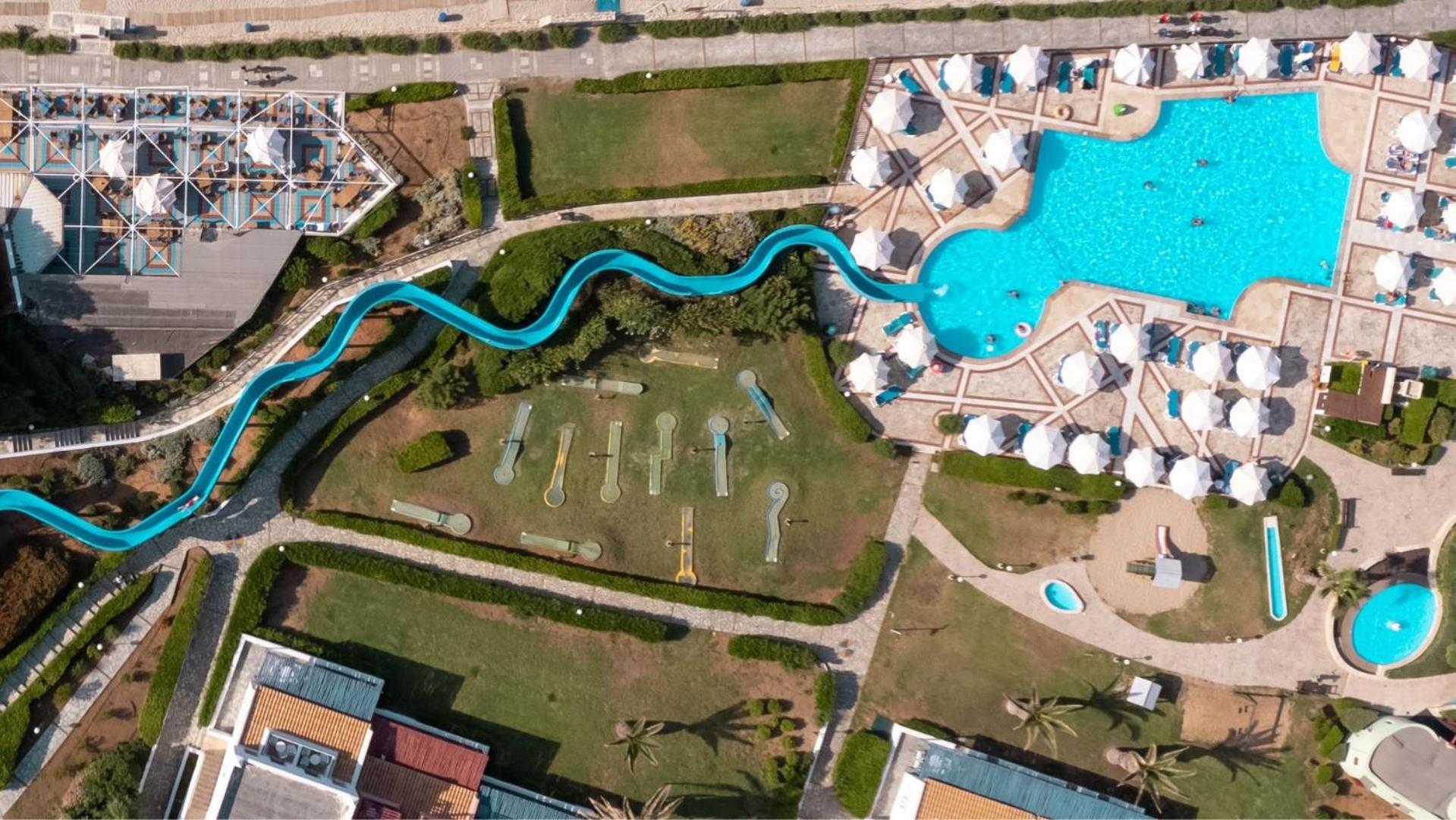Aerofoto skats uz Canvas by Mitsis Cretan Village teritoriju — zils ūdens slidkalniņš, minigolfa laukums un baseins ar saulessargiem