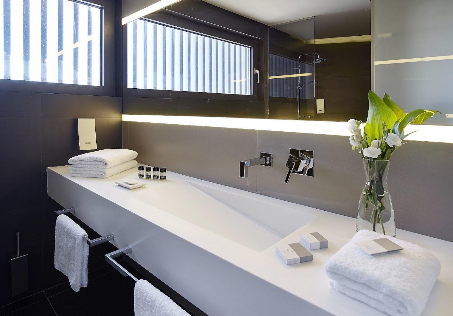 NEMA Design Hotel & Spa moderna vannas istaba ar minimālistisku dizainu, baltu izlietni, lielu spoguli un luksusa kosmētikas piederumiem.