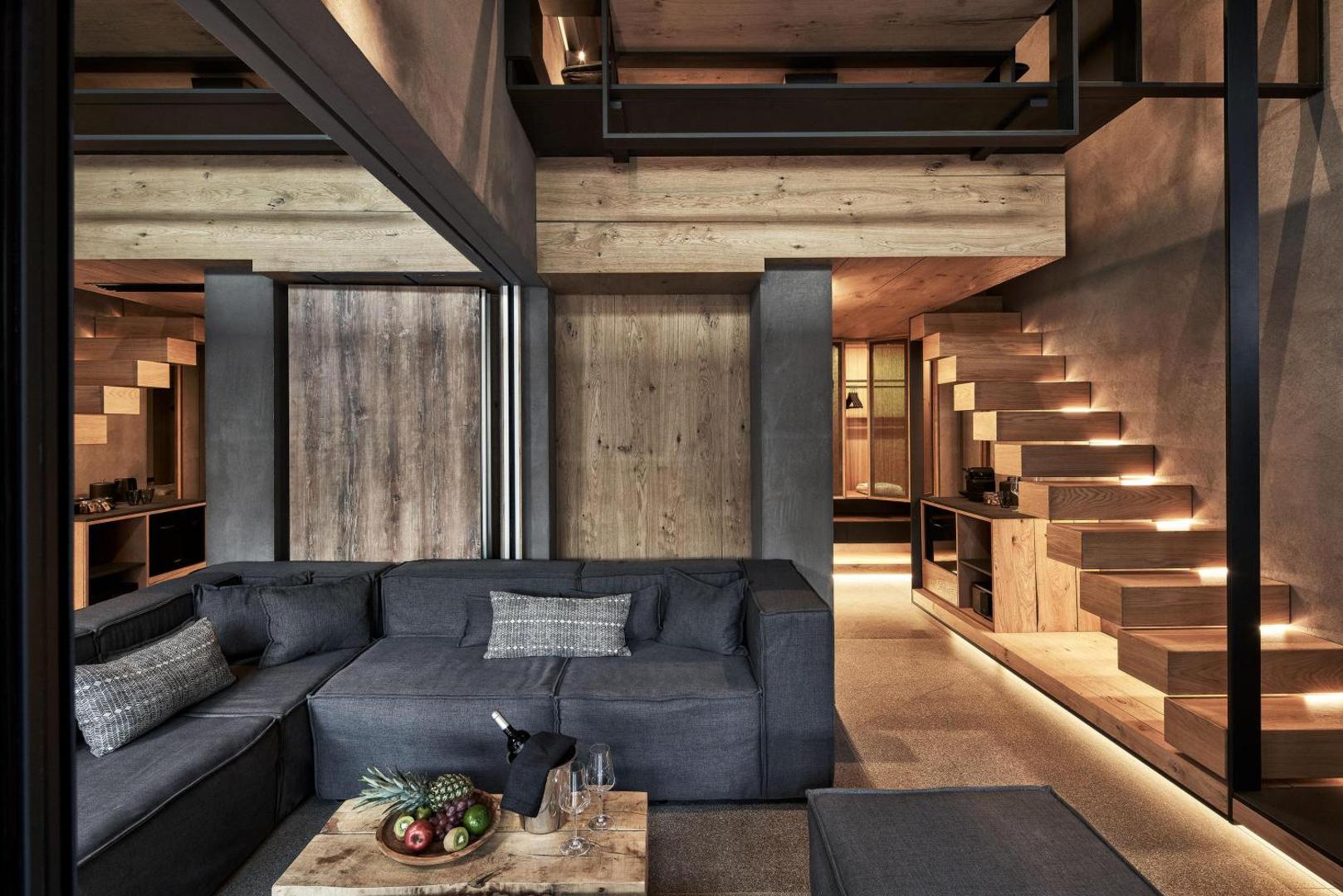 NEMA Design Hotel & Spa divstāvu numura interjers ar tumši pelēku dīvānu, koka kāpnēm un siltu apgaismojumu industriālā stilā.
