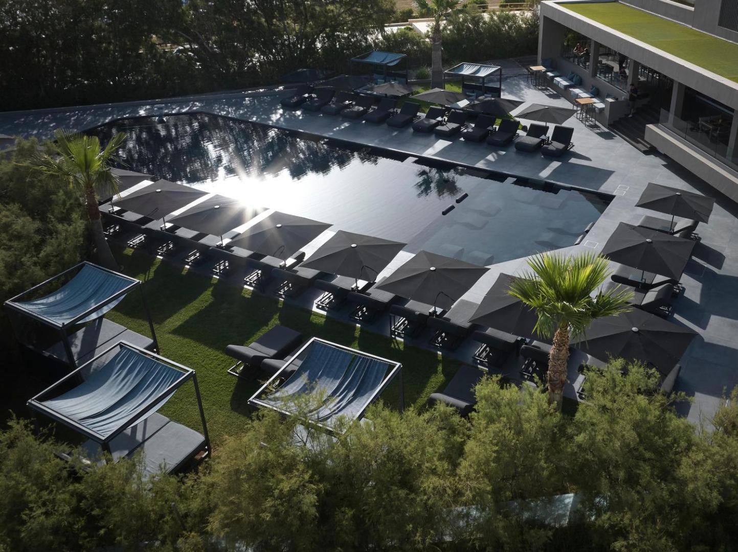 NEMA Design Hotel & Spa galvenais baseins ar melnu apdari, sauļošanās krēsliem un saulessargiem, ko ieskauj zaļi koki un dārzs.