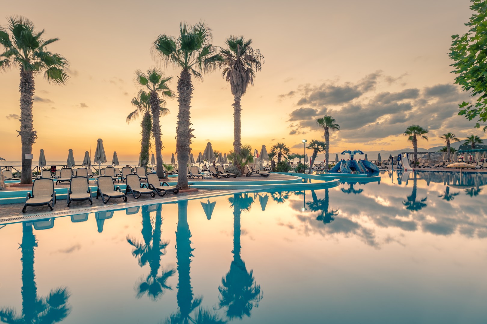 Star Beach Village viesnīcas āra baseins saulrieta laikā ar palmām un atpūtas krēsliem, atspulgs ūdenī Krētas piekrastē