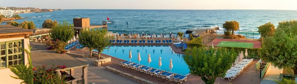 Silva Beach āra baseins ar sauļošanās krēsliem un saulessargiem piekrastē — skats uz Vidusjūru un tenisa kortu fonā