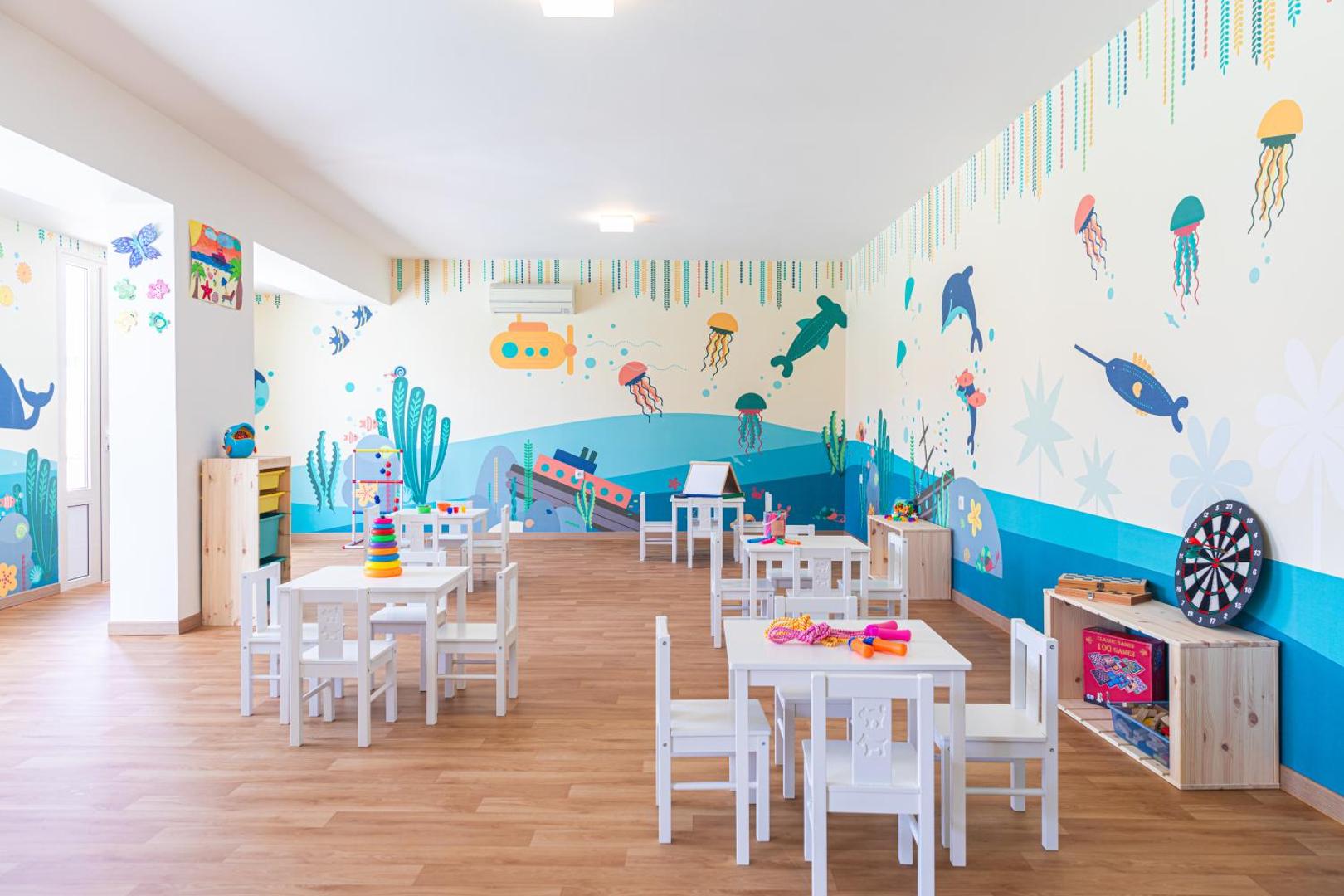 Krāsaina bērnu rotaļu telpa Iberostar Selection Creta Marine viesnīcā ar zīmējumiem uz sienām, maziem galdiņiem un radošām spēlēm