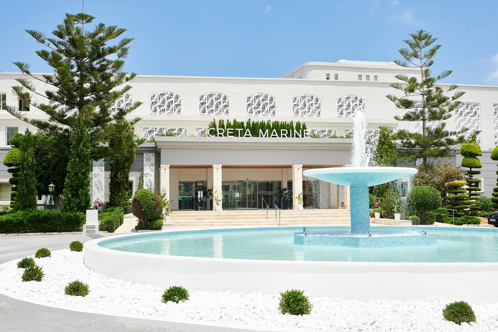 Iberostar Selection Creta Marine galvenā ieeja ar strūklaku, baltu fasādi un koptiem apstādījumiem saulainā dienā.