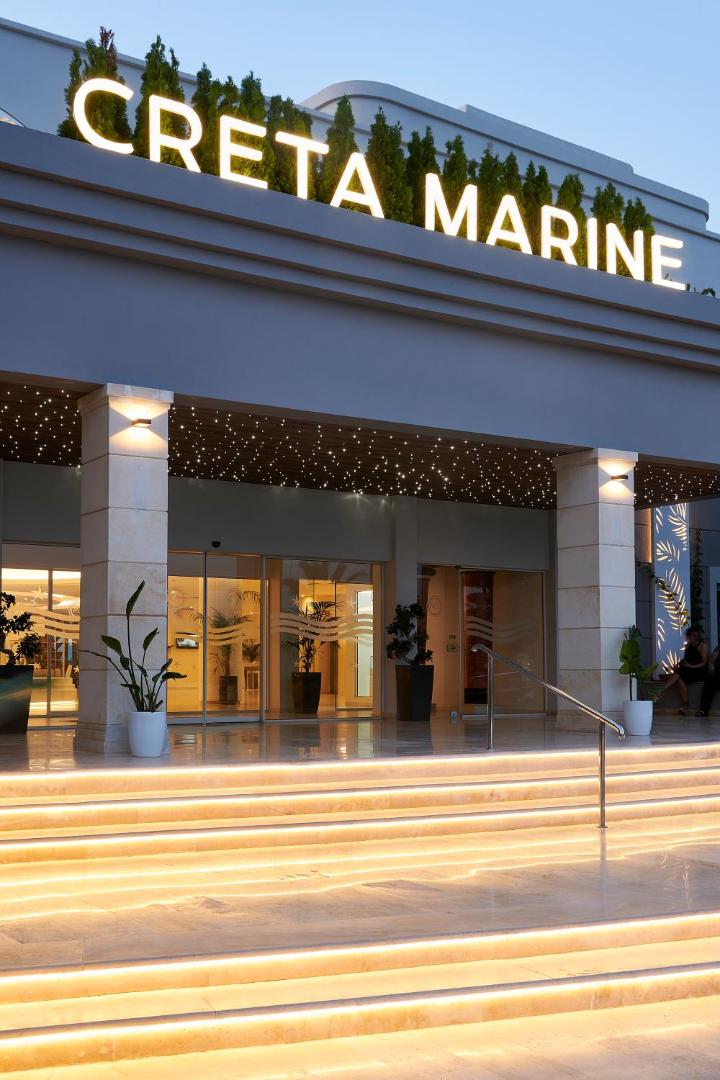 Viesnīcas Iberostar Selection Creta Marine izgaismotā ieeja vakarā ar modernu apgaismojumu un viesnīcas nosaukumu uz fasādes.