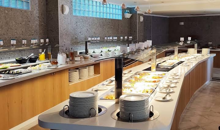 Hydramis Palace Beach Resort restorāna bufetes zona ar plašu siltā ēdiena izvēli un sakārtotiem šķīvjiem uz gaišas letes