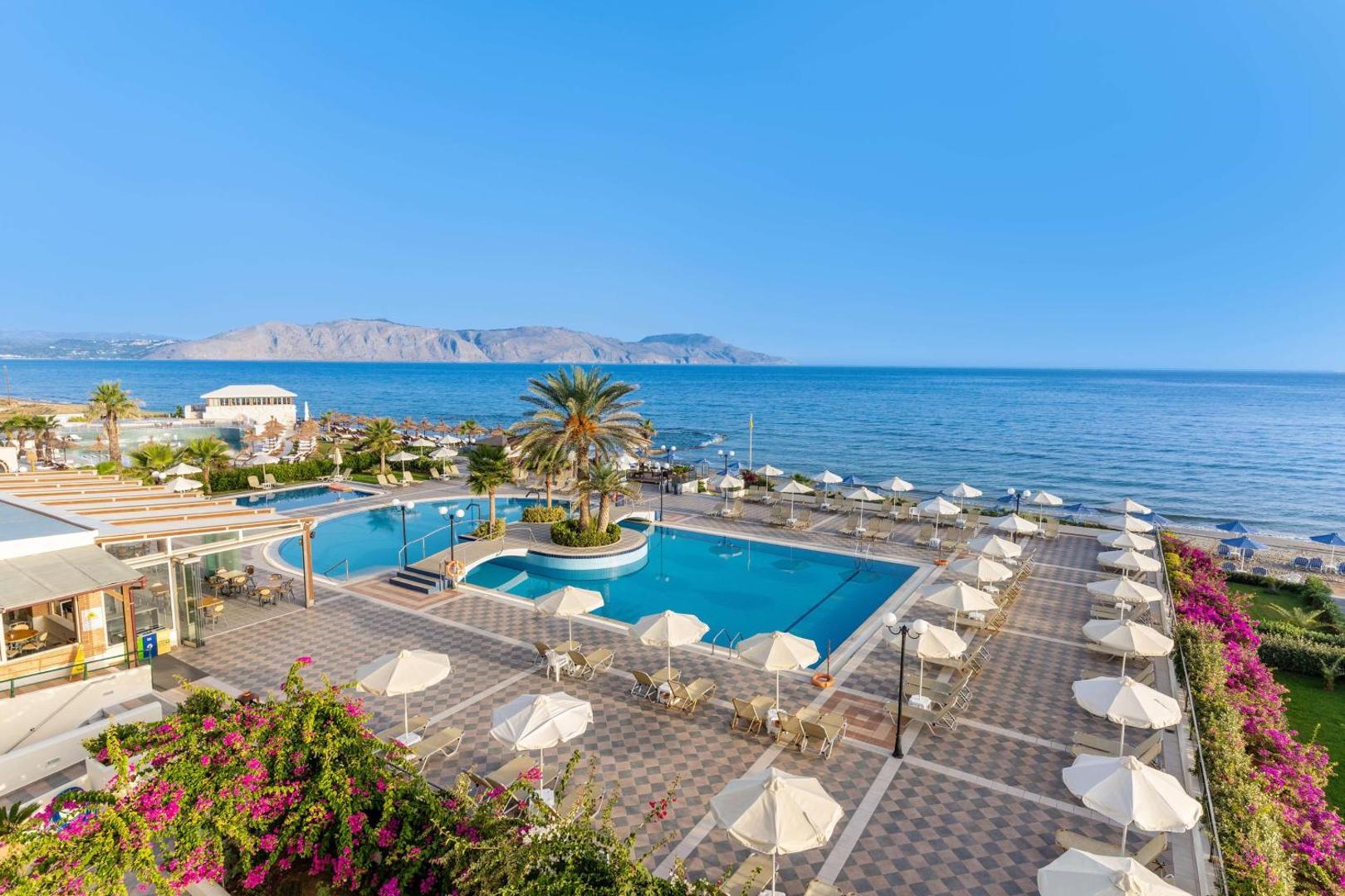 Hydramis Palace Beach Resort baseina zona ar saulessargiem un skatu uz jūru un kalniem saulainā dienā Krētā