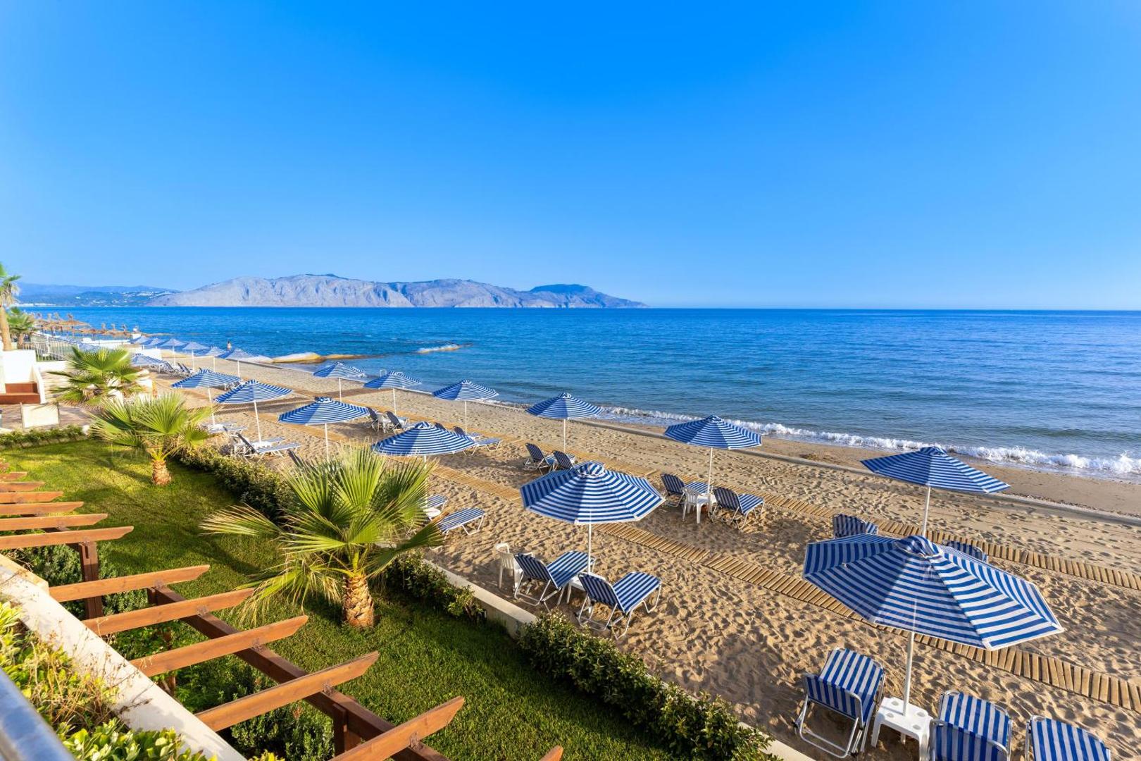 Hydramis Palace Beach Resort plašā smilšu pludmale Krētā ar svītrainiem saulessargiem, sauļošanās krēsliem un skatu uz kalniem tālumā