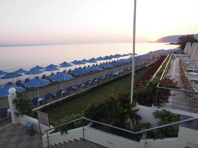 Saulrieta skats no Hydramis Palace Beach Resort terases uz pludmali, kur rindās izkārtoti zili balti saulessargi un sauļošanās krēsli