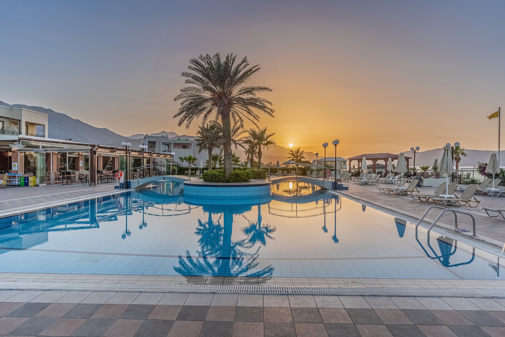 Hydramis Palace Beach Resort āra baseins saulrietā Georgiupoles piekrastē Krētā ar palmām un dekoratīviem tiltiņiem
