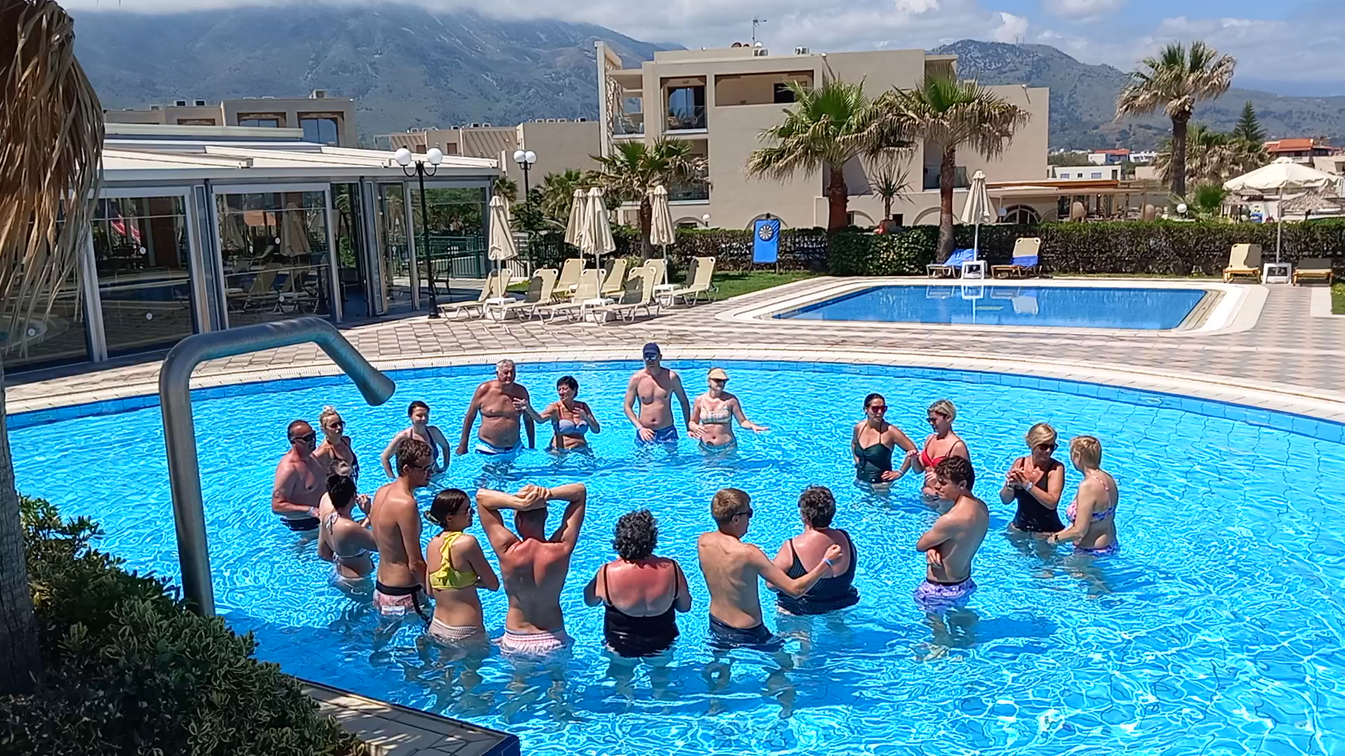 Hydramis Palace Beach Resort āra baseins ar viesiem, kuri piedalās ūdens aerobikas nodarbībā kalnu un palmu fonā Krētā