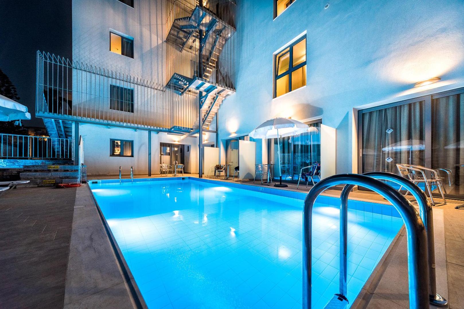 Pollis Hotel izgaismots āra baseins vakarā — moderna arhitektūra, terases ar galdiņiem un mājīgs apgaismojums Hersonisā