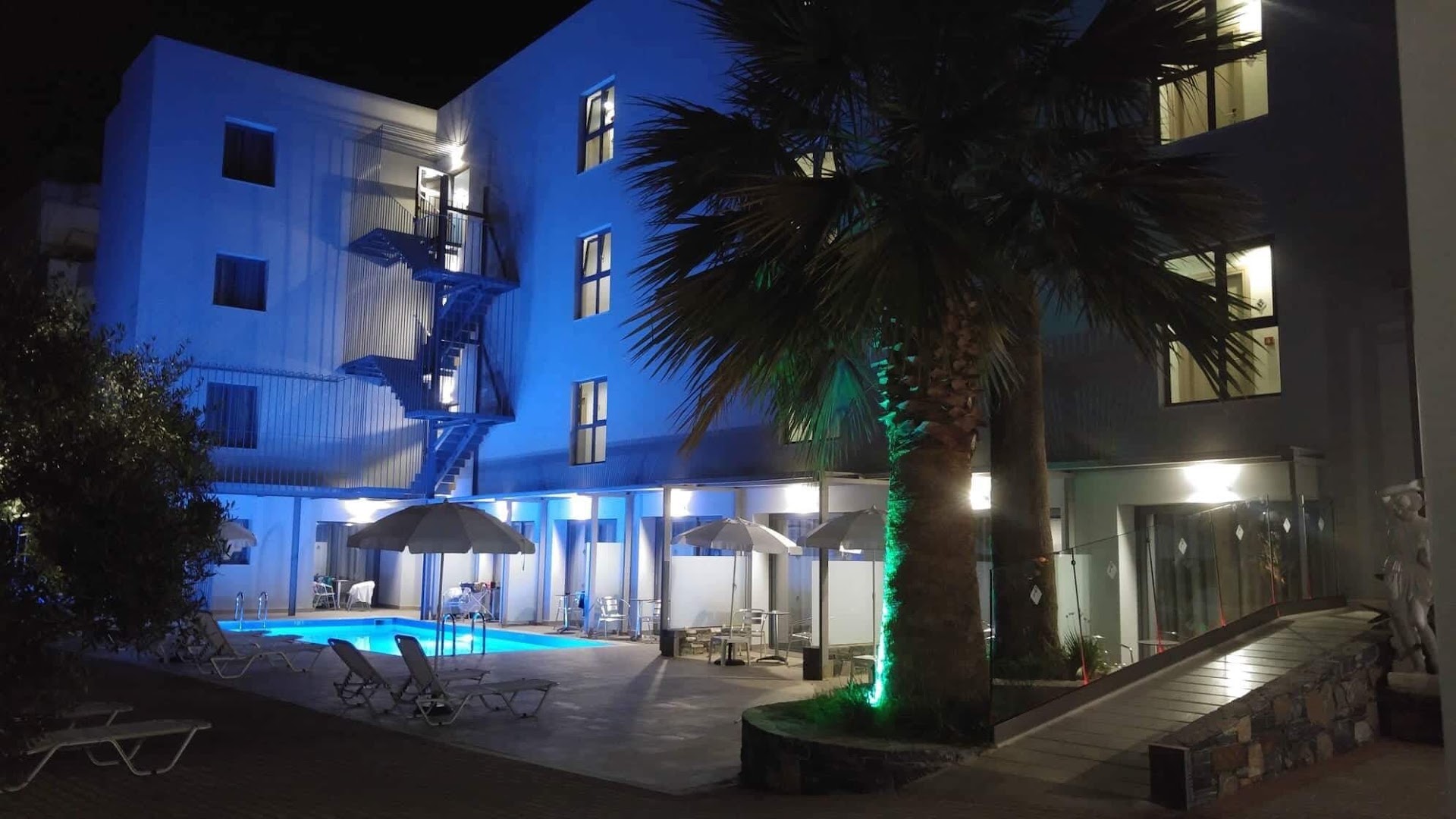 Pollis Hotel iekšpagalms ar āra baseinu naktī — zils apgaismojums, sauļošanās krēsli un saulessargi pie viesnīcas korpusa