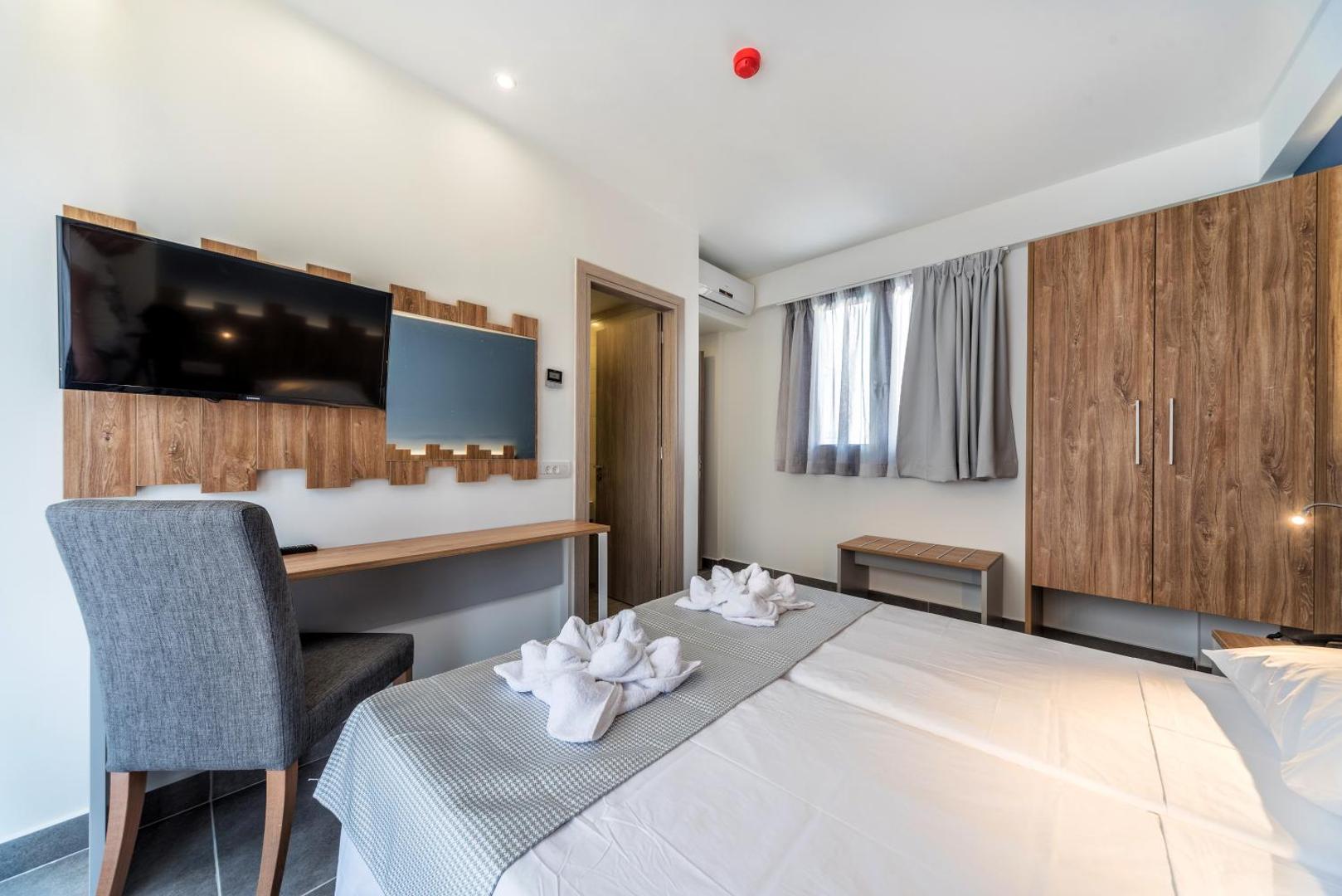 Pollis Hotel numura interjers ar sienas televizoru, rakstāmgaldu un koka skapi — skats uz gultu ar dekoratīviem dvieļiem