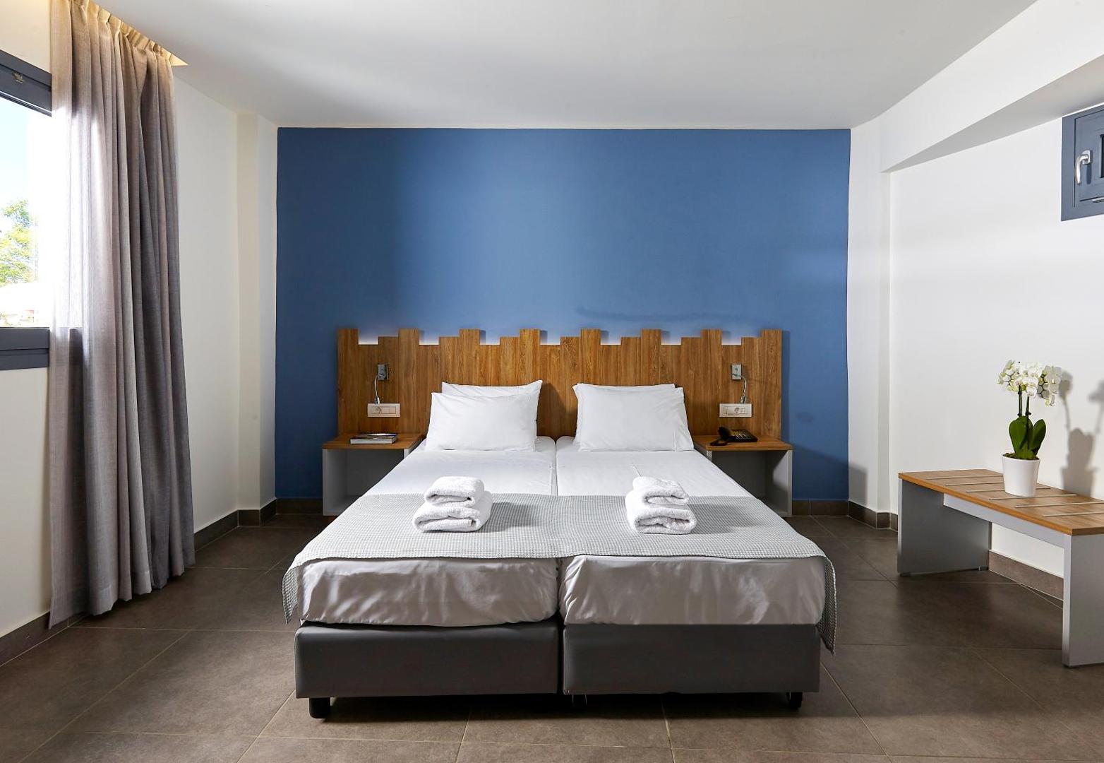 Pollis Hotel gaišs divvietīgs numurs Hersonisā, Krētā — zila akcenta siena, koka galvgalis un divas gultas ar baltu veļu