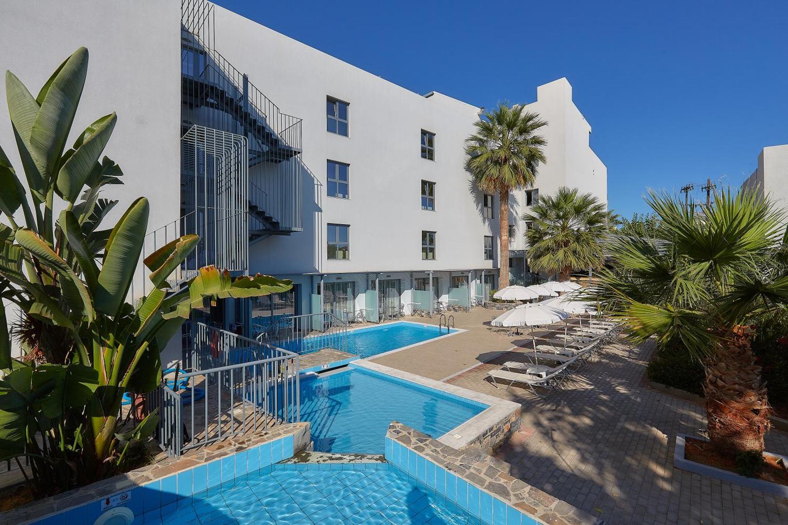 Pollis Hotel āra baseins Hersonisā, Krētā — moderna balta ēka, sauļošanās krēsli ar saulessargiem un palmas pie baseina zonas