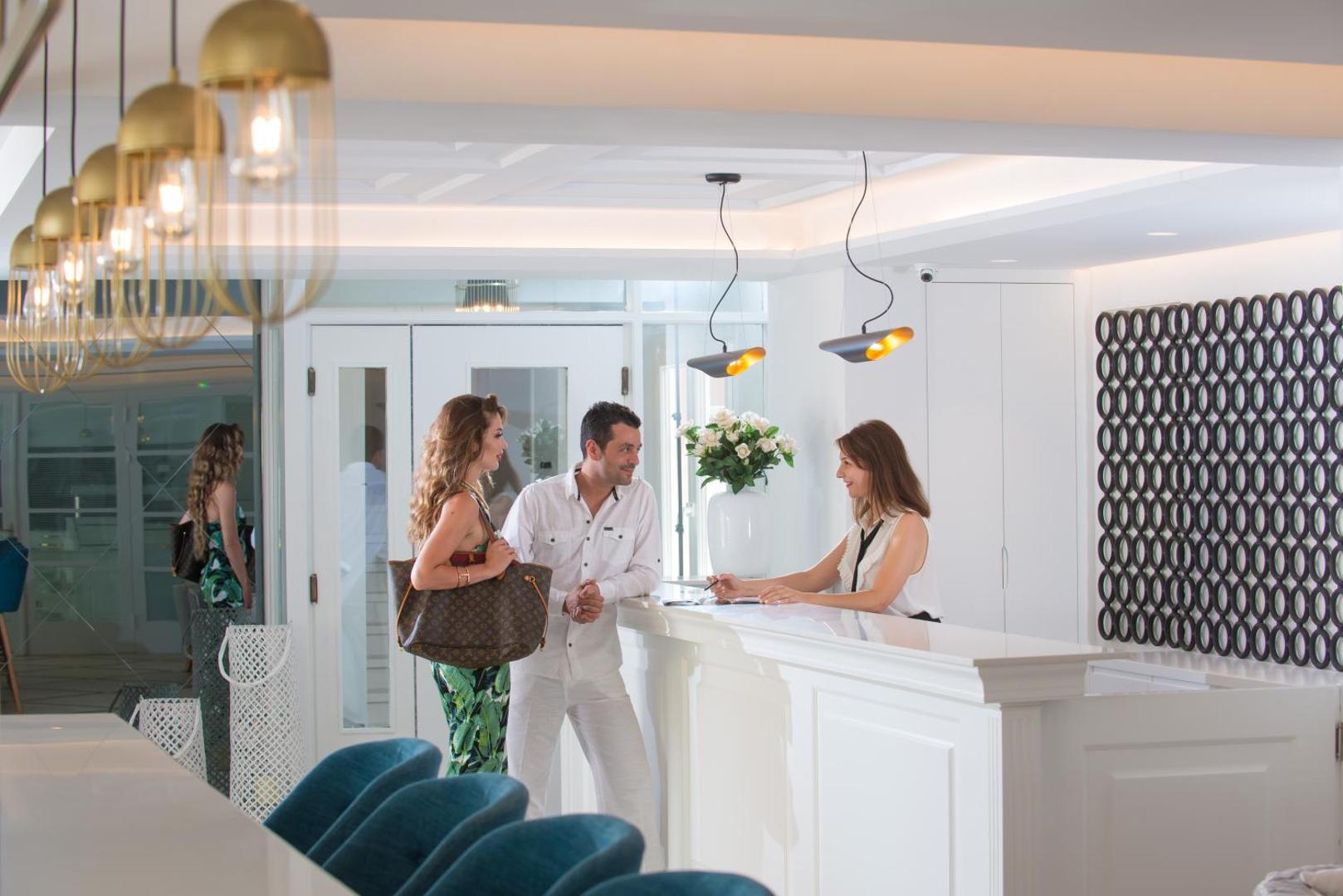 Mari Kristin Beach Hotel reģistratūras zona ar viesmīlīgu darbinieci un viesiem pie baltas letes modernā lobby interjerā